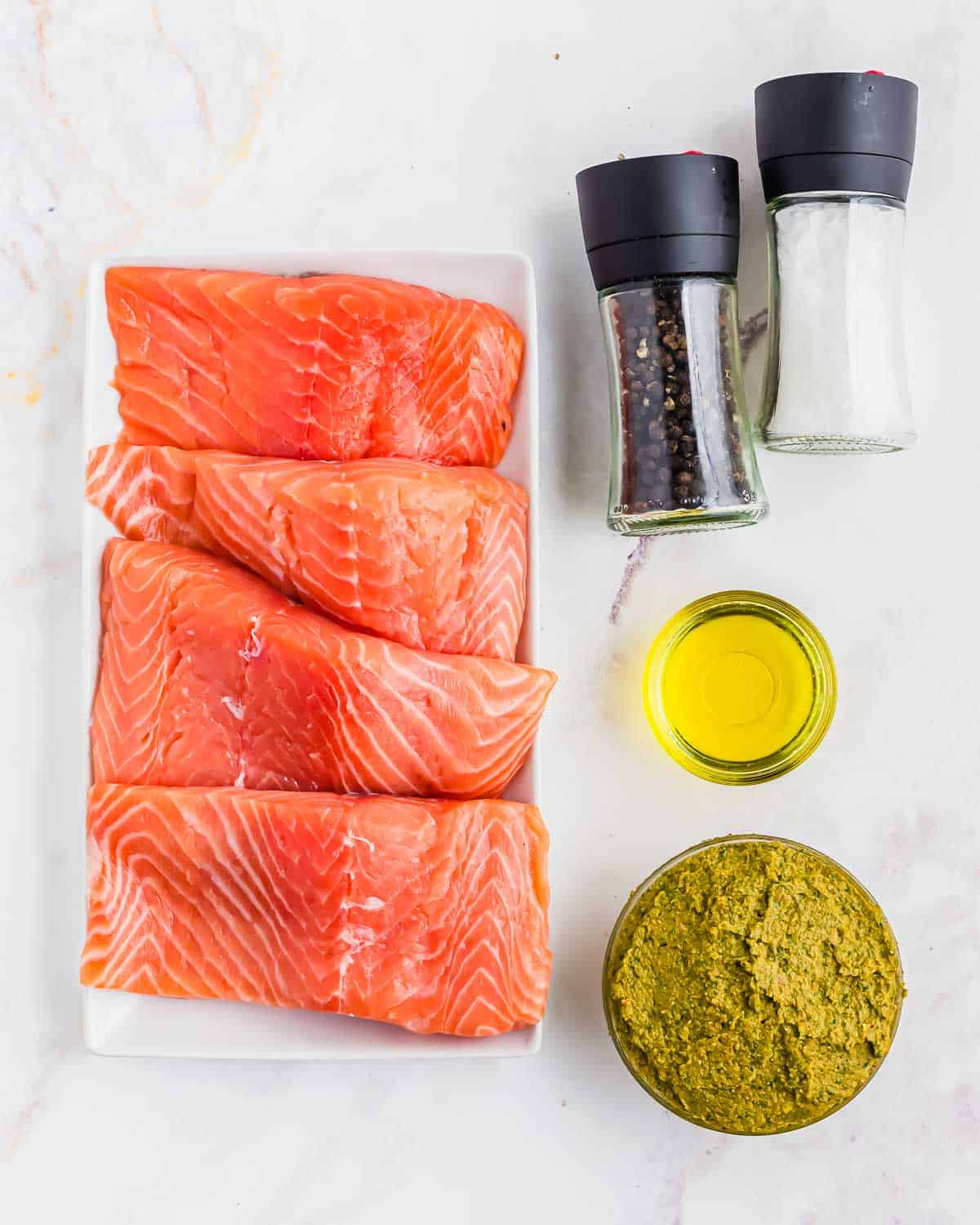 A plate of raw salmon with a small bowl of pesto, salt and pepper shakers and olive oil.