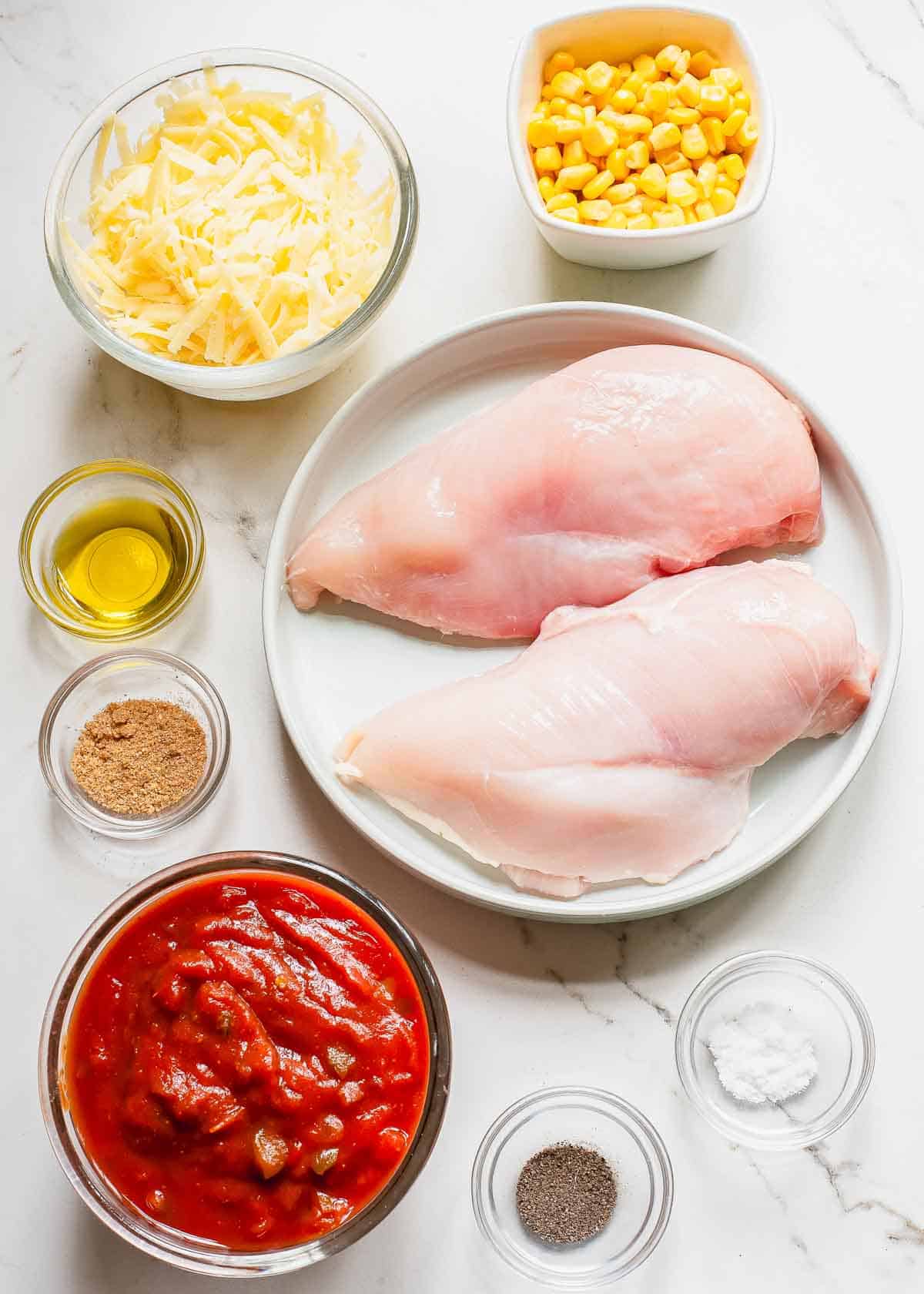 Chicken breasts, corn, tomatoes and other ingredients on a white marble table.