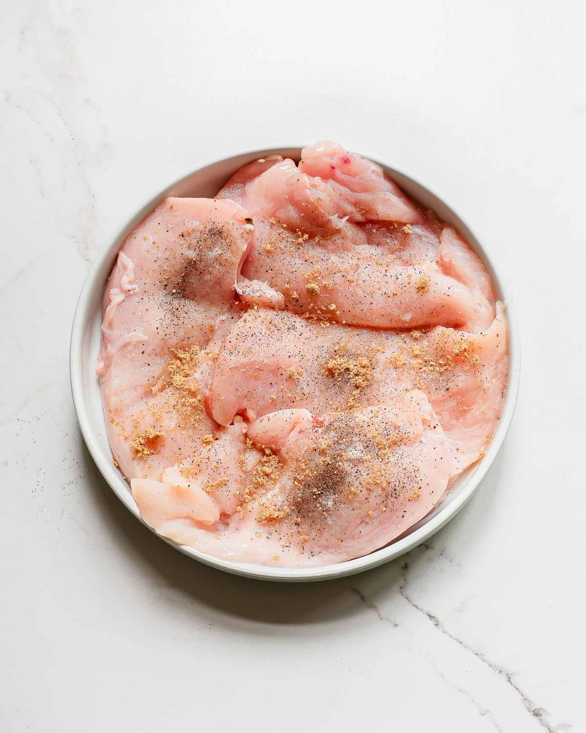 Chicken breasts in a plate on a marble countertop seasoned with taco seasoning, salt and pepper.