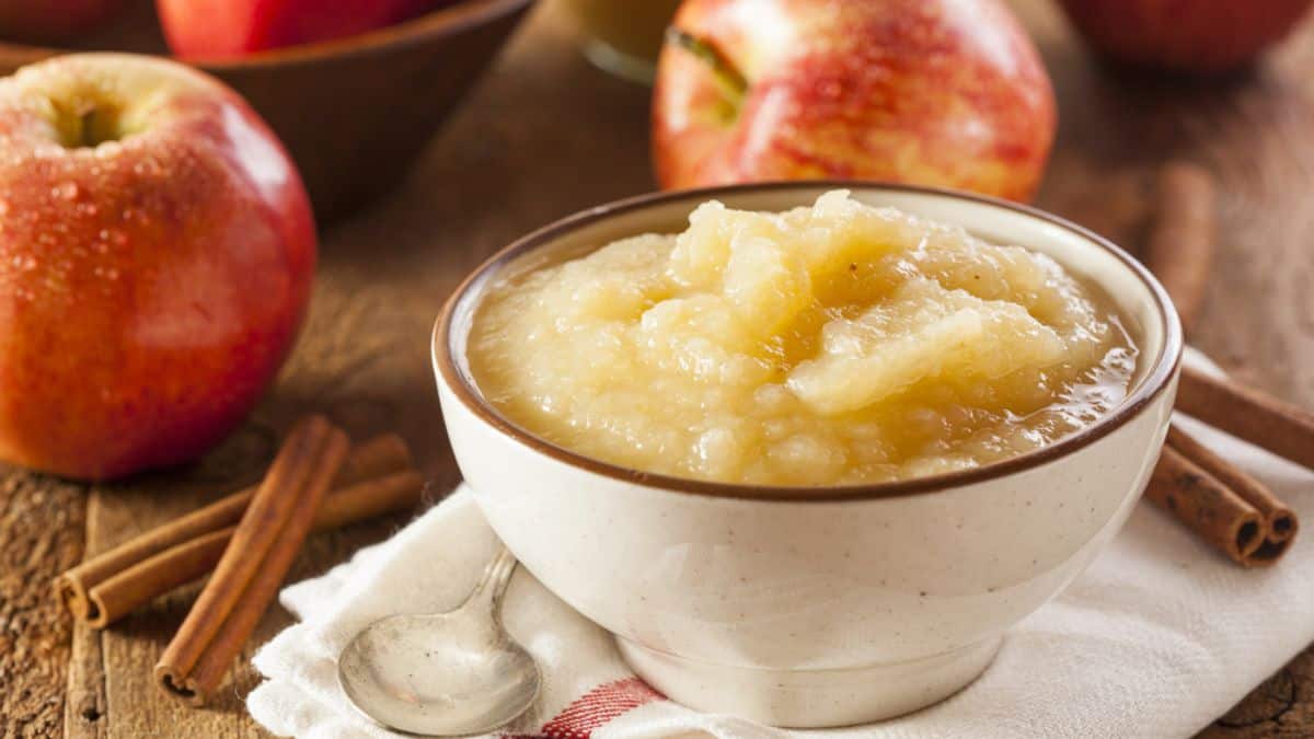 A bowl of homemade applesauce with cinnamon sticks and fresh apples in the background.
