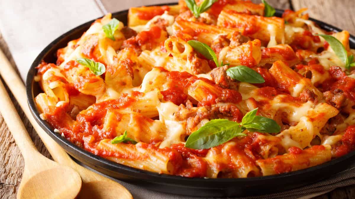 A skillet of baked ziti pasta with tomato sauce, melted cheese, and fresh basil leaves.