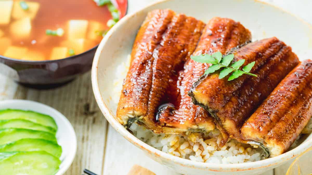 A bowl of unagi don (grilled eel over rice) garnished with a sprig of herbs, accompanied by slices of cucumber and a bowl of soup.