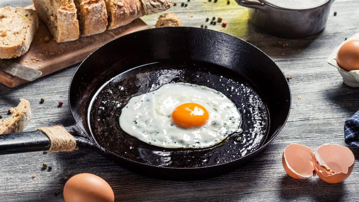 A single fried egg in a cast iron skillet surrounded by ingredients and utensils on a wooden table.