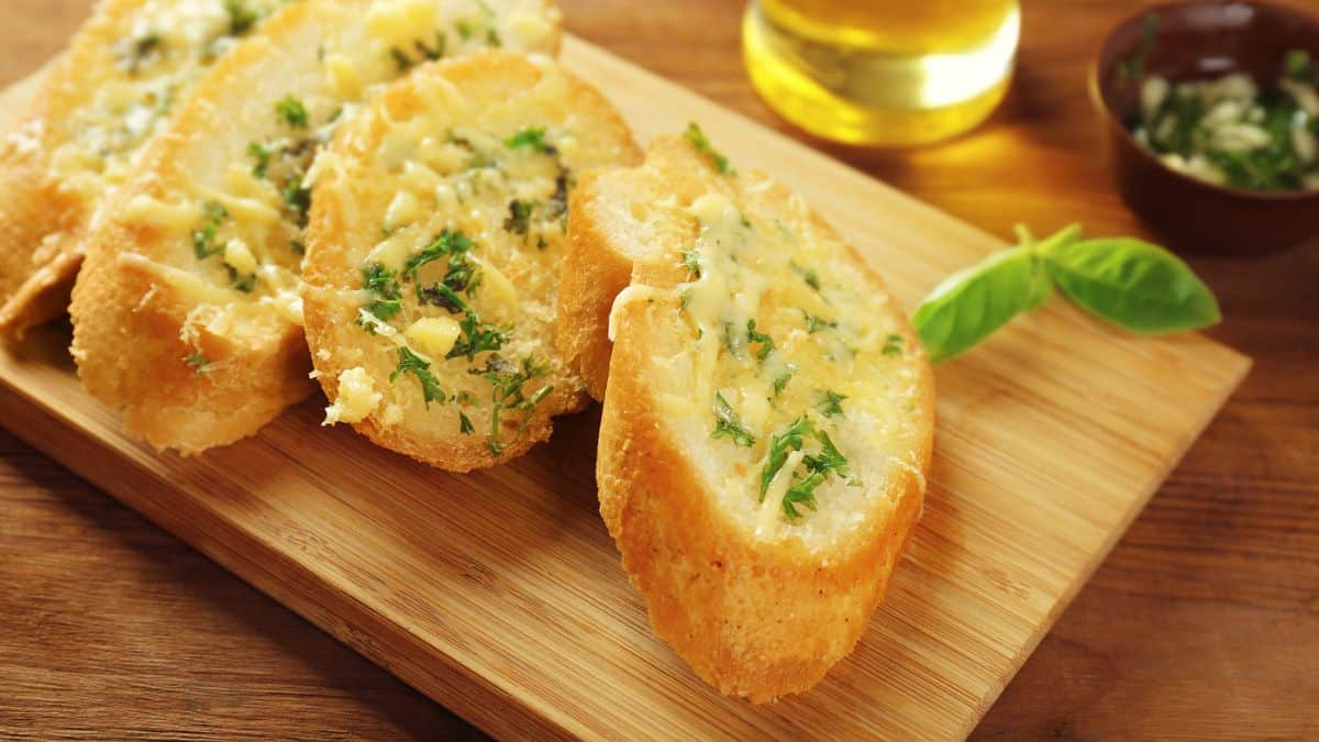 Slices of garlic bread with melted cheese and herbs on a wooden cutting board.