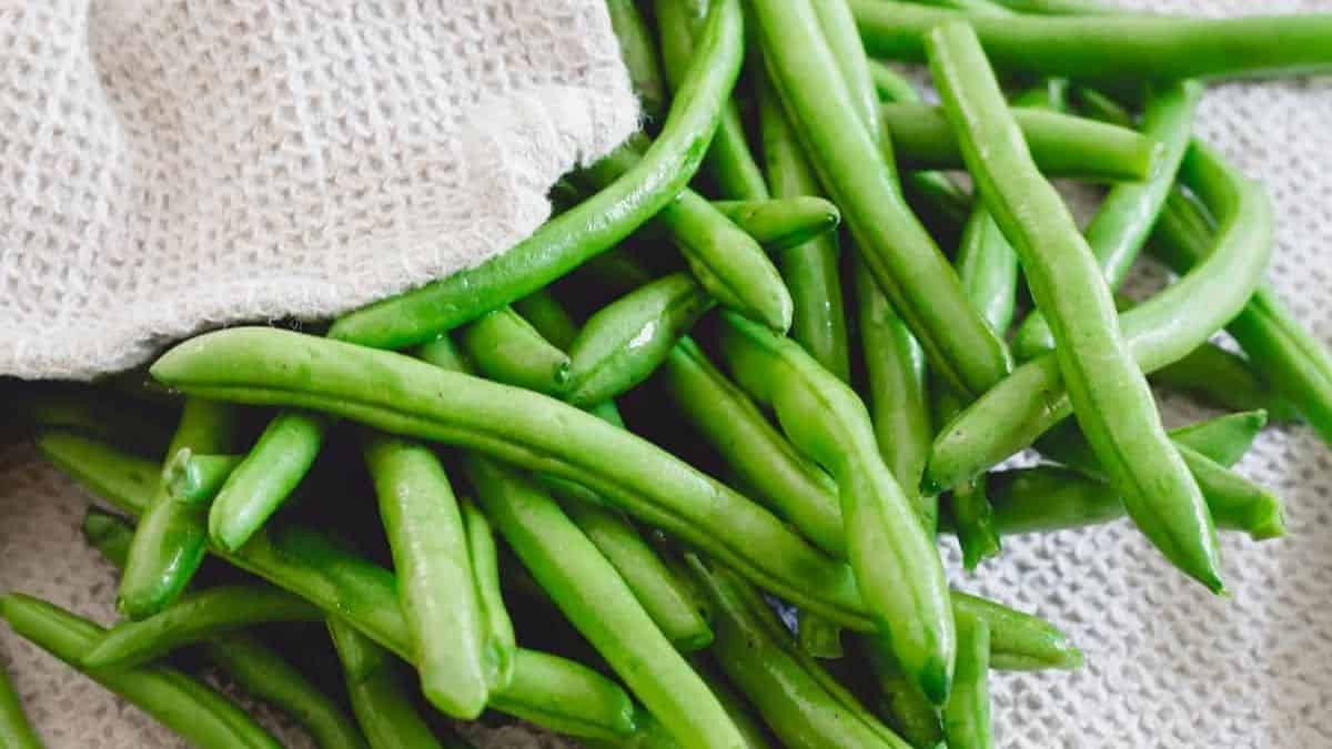 Fresh green beans spilling out from under a beige fabric hat onto a textured surface.