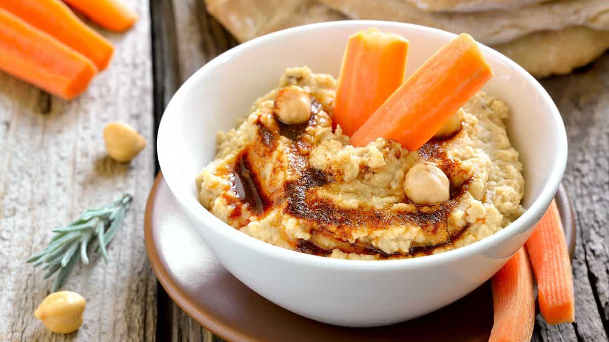 A bowl of hummus garnished with paprika and whole chickpeas, accompanied by carrot sticks on a wooden surface.