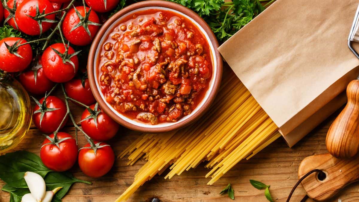 A bowl of meaty tomato sauce beside fresh ingredients and uncooked spaghetti.