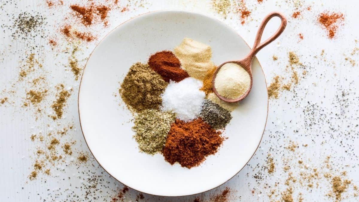 A variety of colorful spices and seasonings arranged neatly on a white plate with a wooden spoon, ready to add flavor to any food you make yourself.
