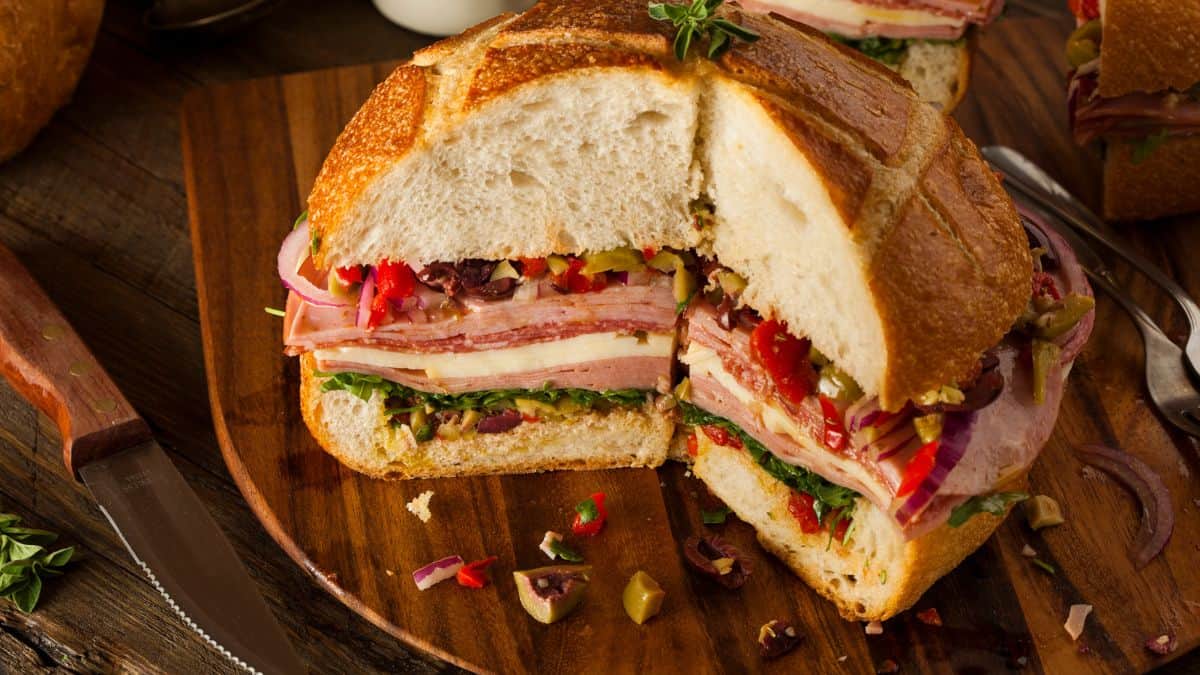 A stacked muffuletta sandwich on a wooden board with meats, cheese, and olive salad.