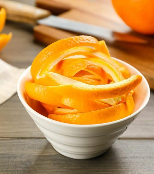 A white bowl filled with orange peels on a wooden surface with whole oranges in the background.