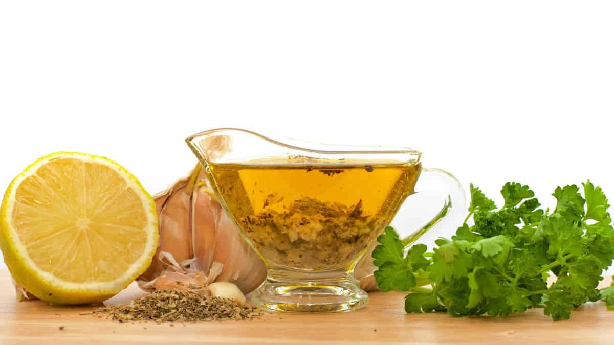 A glass gravy boat with herb-infused oil, accompanied by a garlic bulb, half a lemon, and fresh parsley on a wooden surface against a white background.