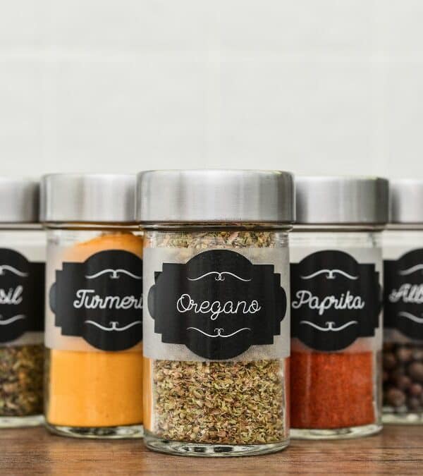 Five spice jars with labeled contents including basil, turmeric, oregano, paprika, and allspice on a kitchen counter.