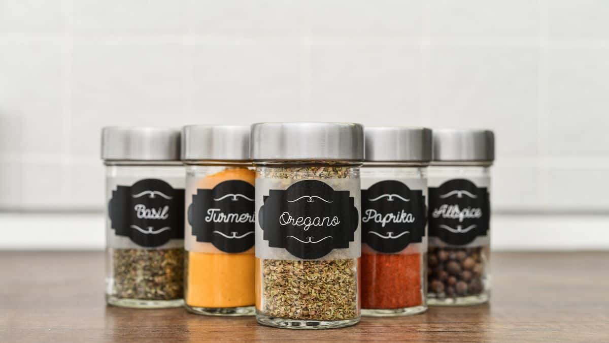Five spice jars with labeled contents including basil, turmeric, oregano, paprika, and allspice on a kitchen counter.