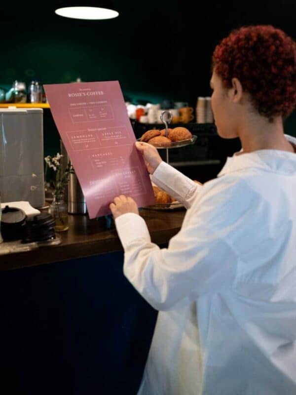 A person in a white shirt reading a pink menu beside a display of muffins at a counter.