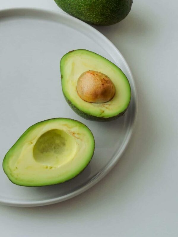 Halved avocado with pit on a white plate, whole avocado in the background.