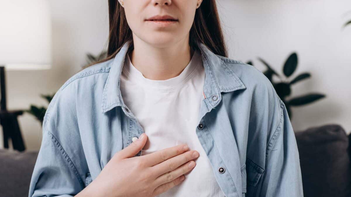 Woman in a white shirt and denim jacket with her hand on her chest.