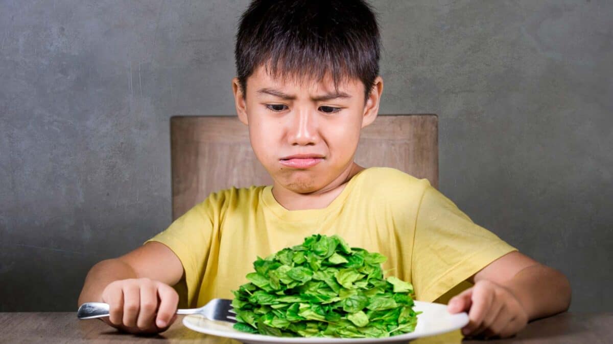 A child appears displeased while looking at a plate full of leafy greens.