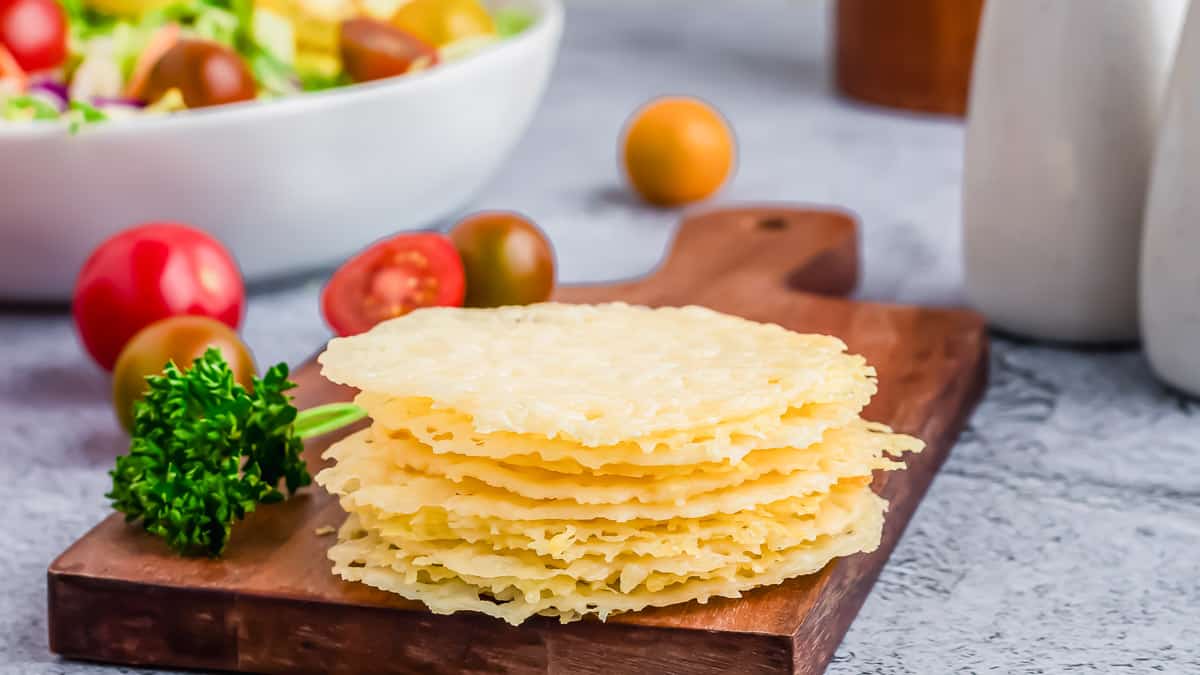 A wooden cutting board with parmesan crisps stacked in a pile.