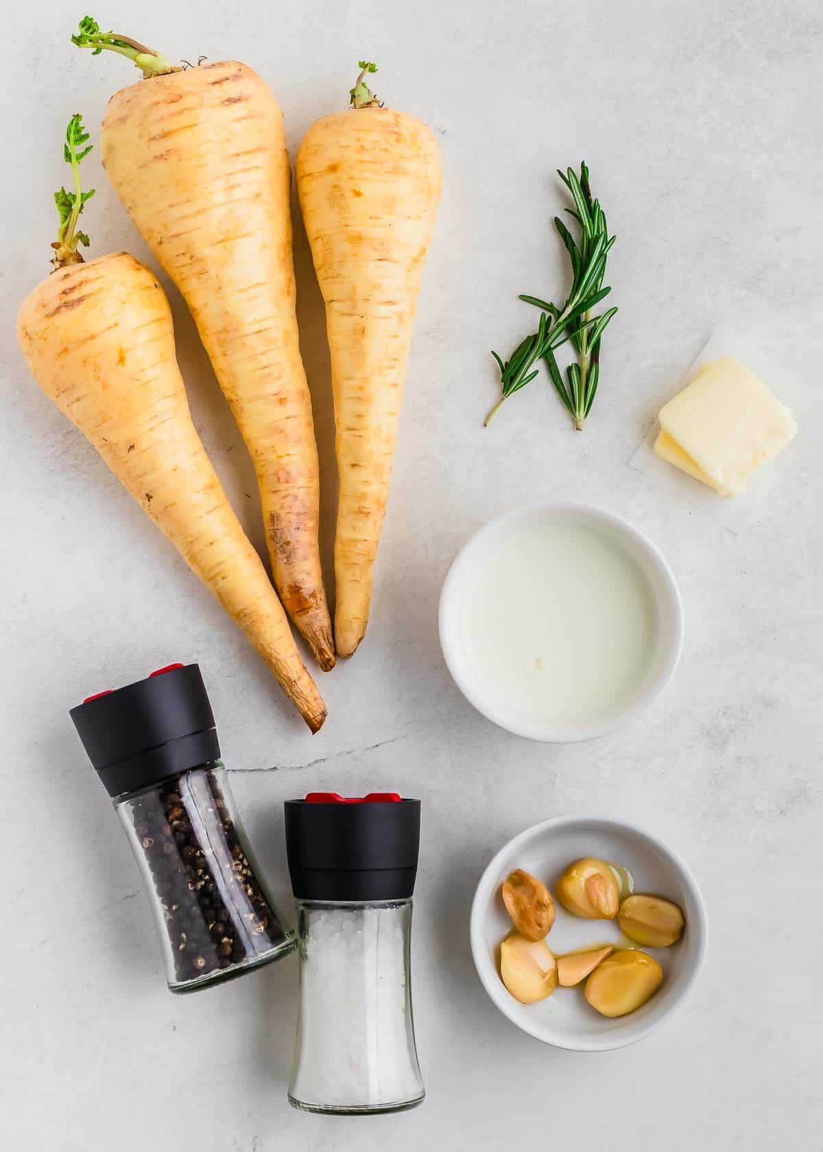 Fresh ingredients for cooking parsnips, herbs, garlic, butter, milk, salt, and pepper on a kitchen countertop.