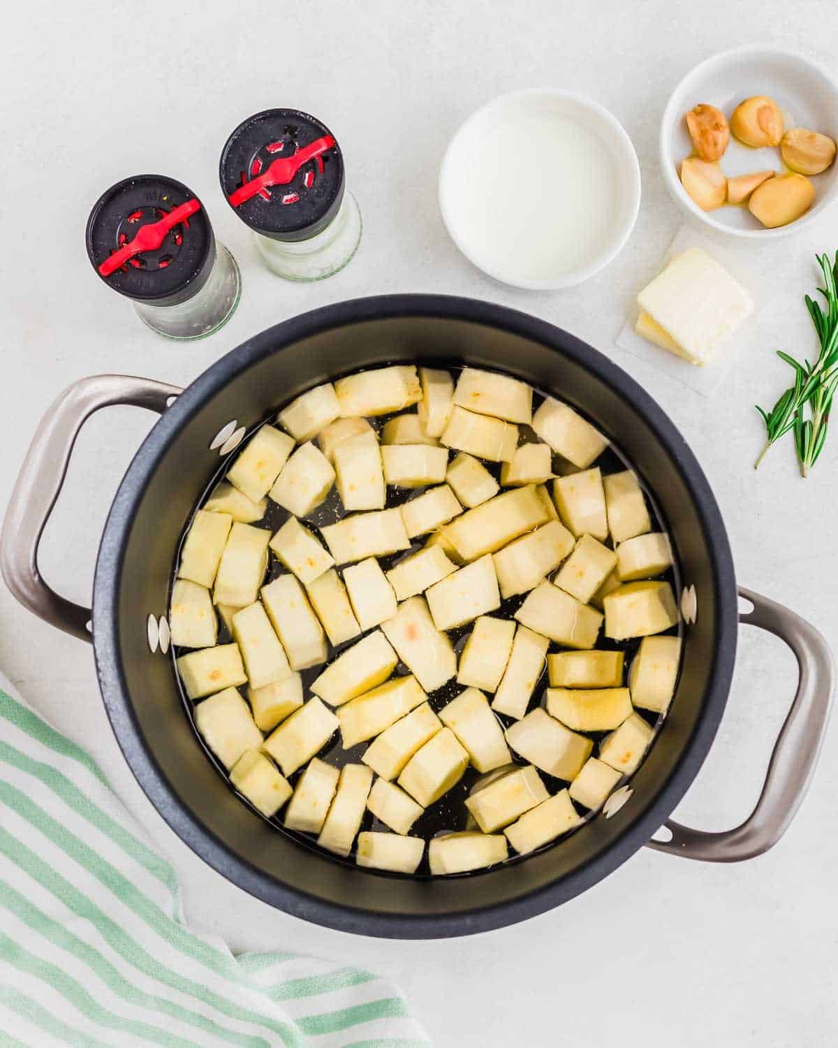Chopped parsnips in a pot with ingredients like milk, butter, and rosemary on the side, prepared for cooking.