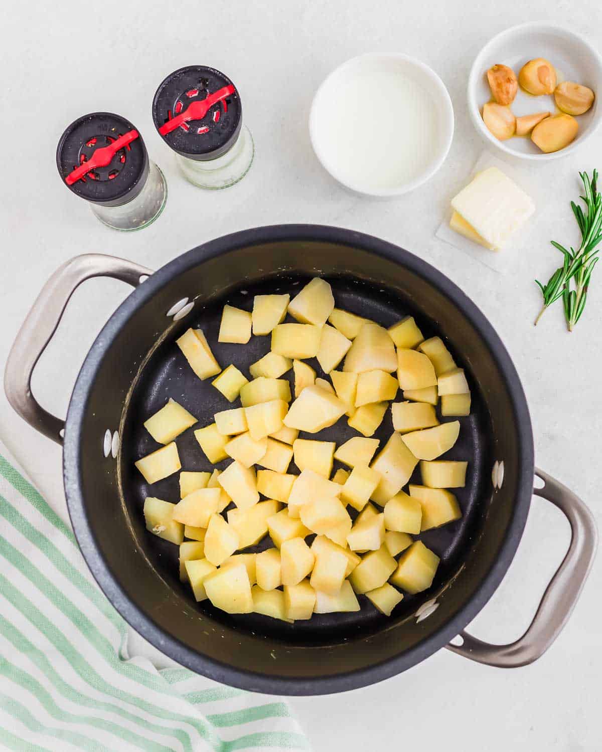 Boiled parnsips in a pot with small bowls of garlic, milk, butter, rosemary, salt and pepper on the side.