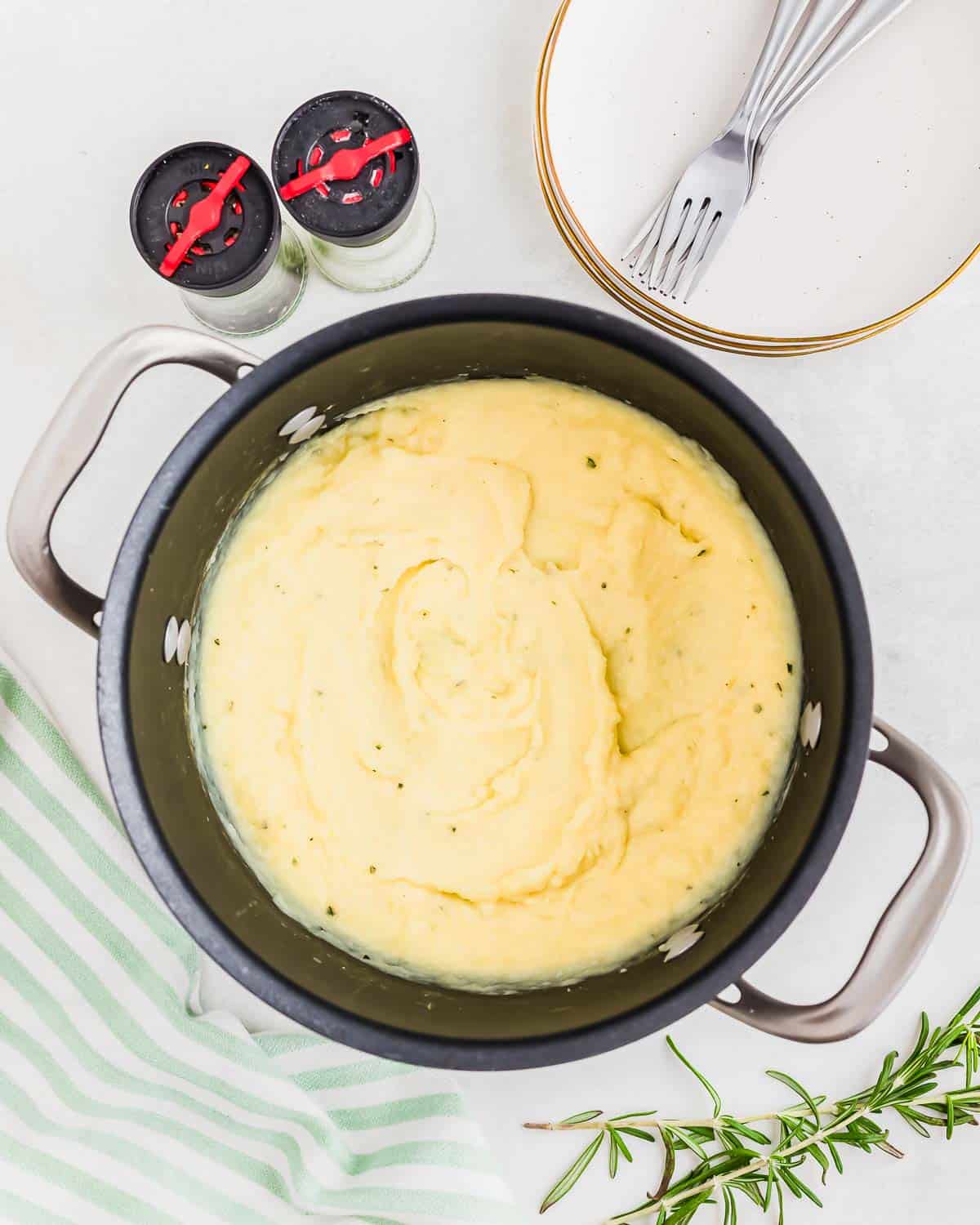 A pot of creamy mashed parsnips on a striped napkin with rosemary sprigs, a plate, forks, and salt and pepper shakers nearby.