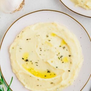 Plate of creamy mashed parsnips garnished with herbs, with garlic and rosemary in the background.