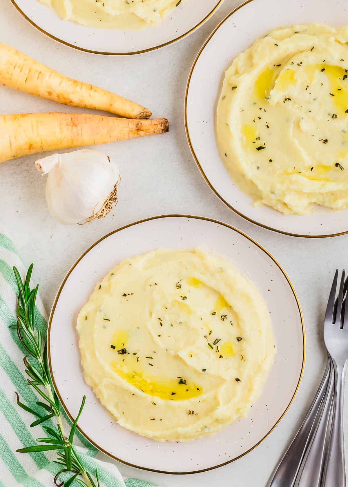 A plate of creamy parsnip puree garnished with herbs, accompanied by whole parsnips, garlic, and rosemary, arranged on a light surface.