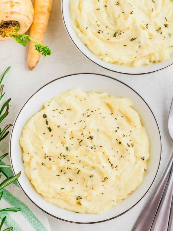 A bowl of creamy mashed parsnips garnished with herbs, accompanied by fresh ingredients and cutlery on the side.