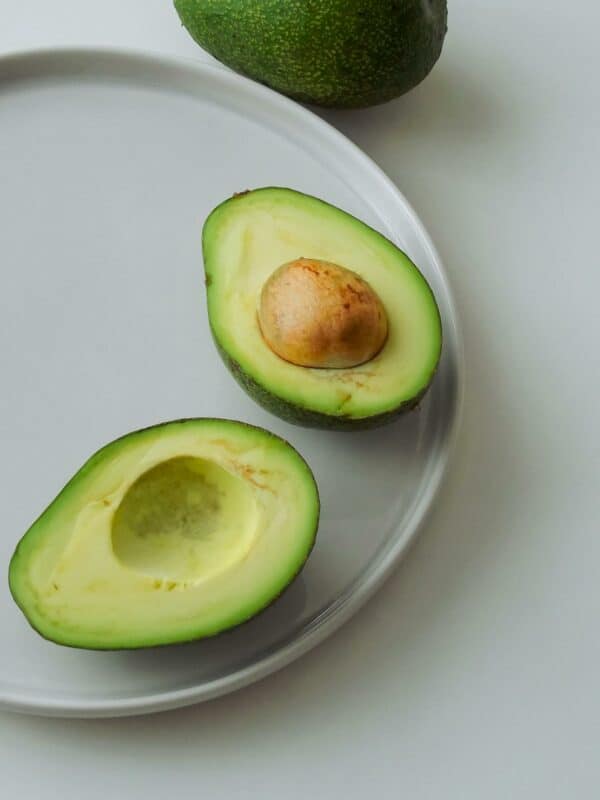 A halved avocado with its stone on a white plate, with a whole avocado in the background.