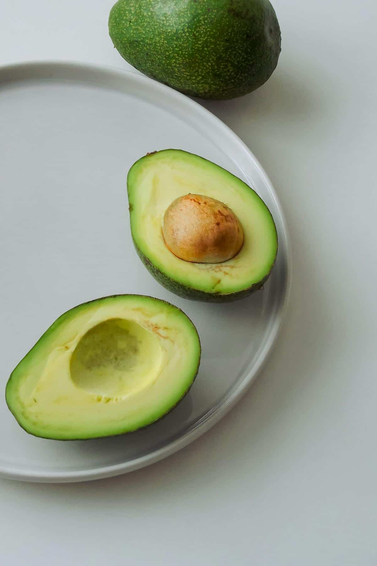A halved avocado with its stone on a white plate, with a whole avocado in the background.