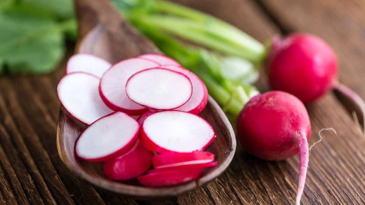 Freshly sliced radishes on a wooden spoon beside whole radishes on a rustic table.