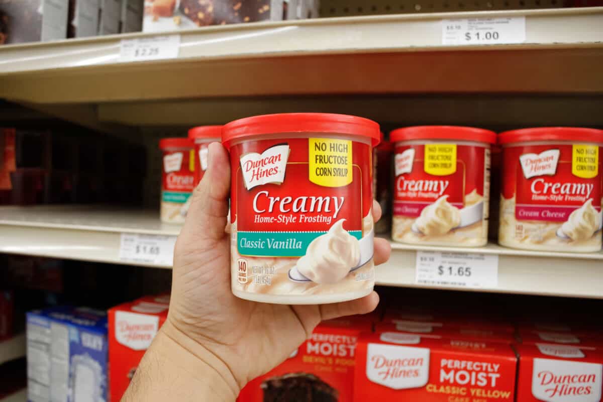 A hand holding a container of duncan hines creamy home-style classic vanilla frosting in a grocery store aisle.