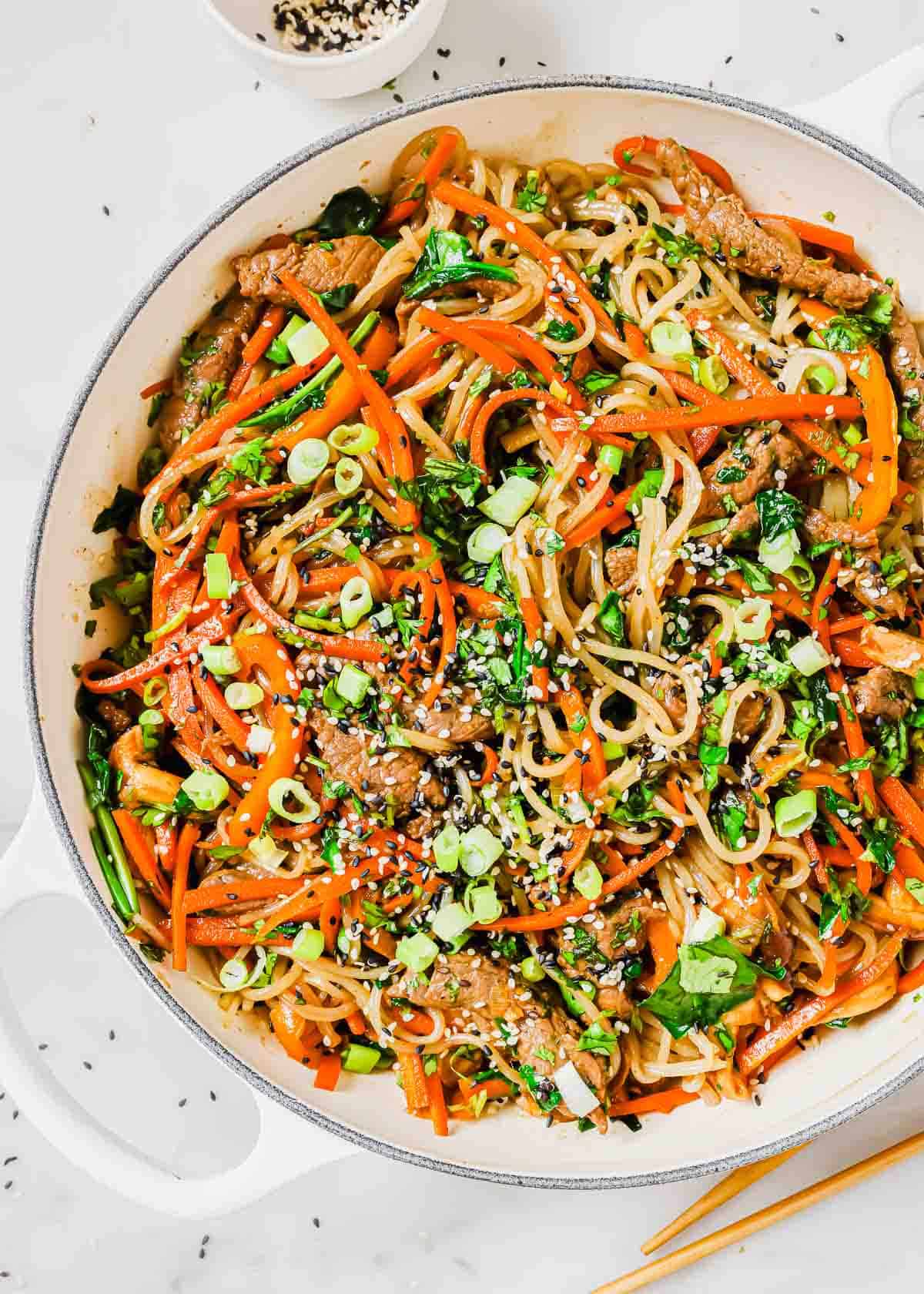 A skillet full of Korean sweet potato noodles and vegetables with chopsticks.