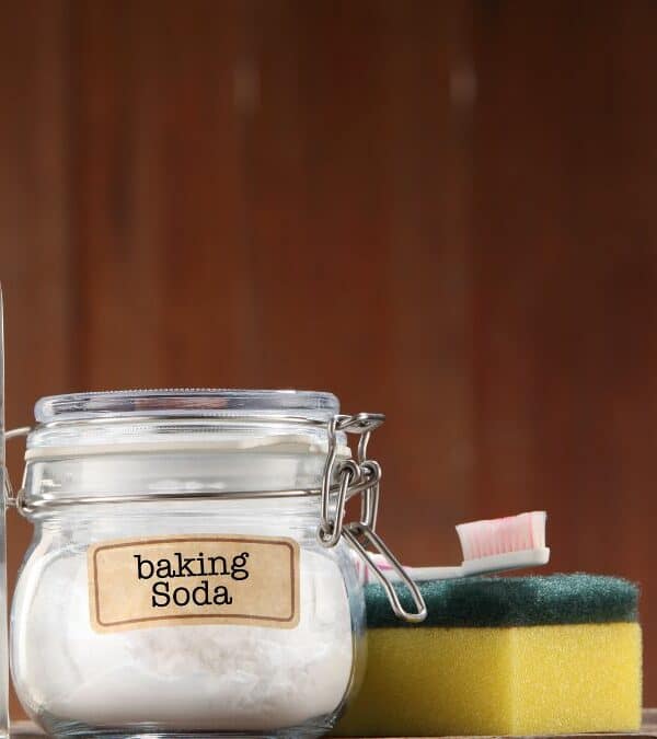 A bottle of vinegar and a jar of baking soda labeled respectively, positioned next to a sponge and scrub brush on a wooden surface.