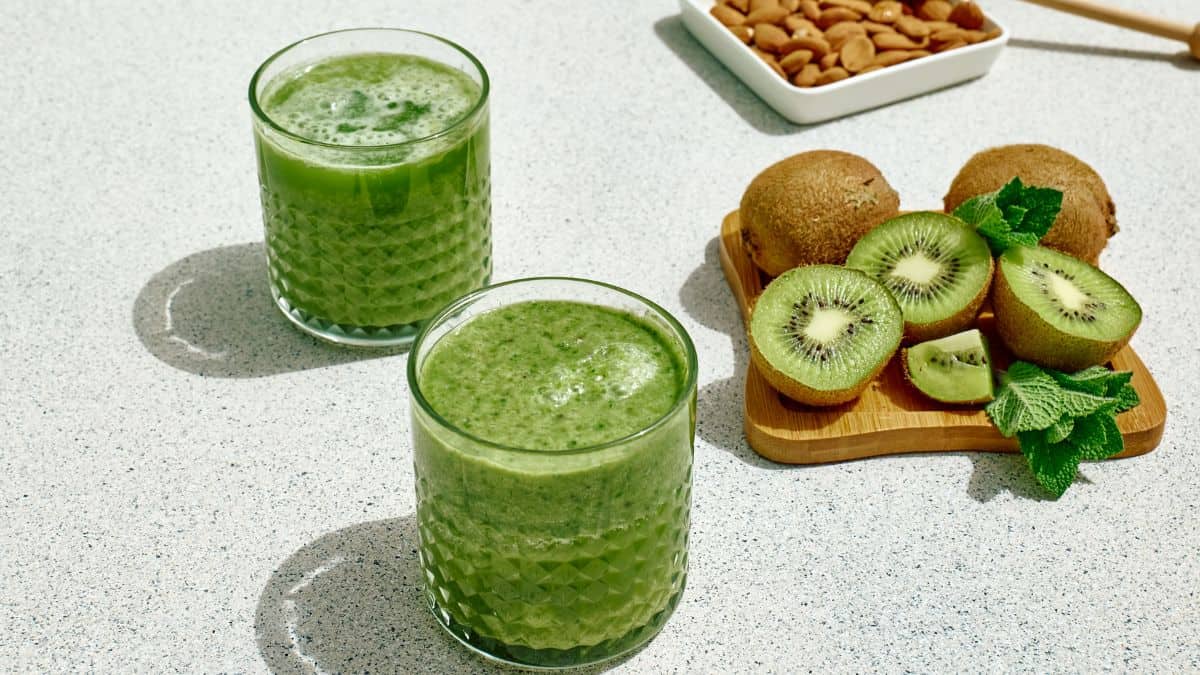 Two glasses of green smoothie on a sunlit table, accompanied by kiwi slices, whole kiwis, fresh mint, and a bowl of almonds.