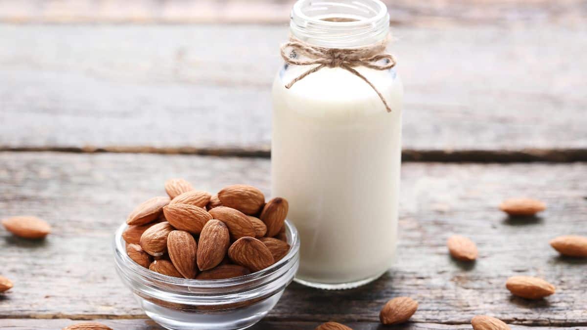 A glass bottle of almond milk tied with twine next to a bowl of whole almonds on a wooden surface.