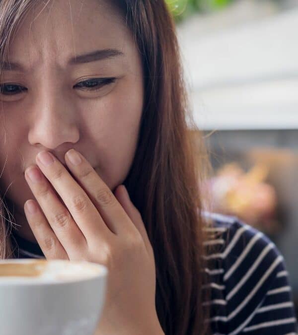 Woman covering her mouth with her hand, holding a cup of coffee, appearing to be coughing or clearing her throat.