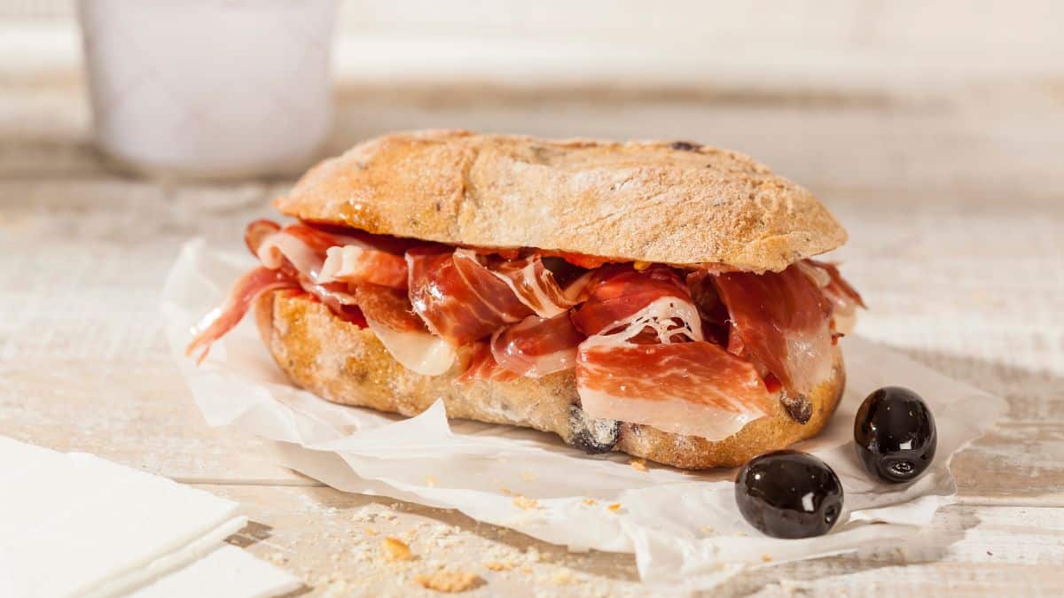 A ciabatta sandwich filled with cured ham and accompanied by black olives on a wooden surface.
