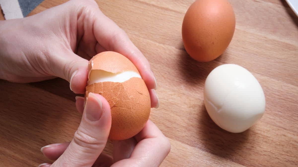 A person peeling a hard-boiled egg over a wooden table, with another unpeeled egg and a peeled egg beside them.