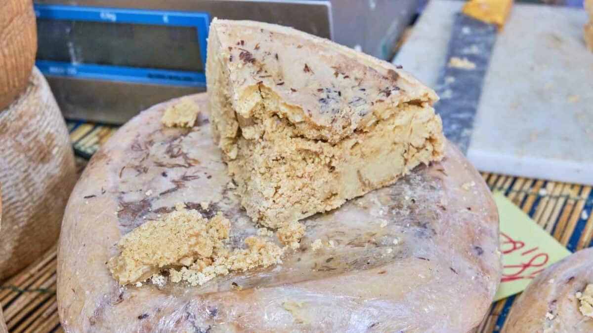 A large wheel of cheese with nuts, partially cut, displaying a crumbly texture, placed on a table at a market stall.