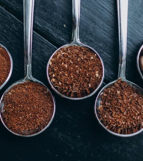 Five spoons containing various stages of coffee preparation, from ground to whole beans, arranged on a wooden surface.