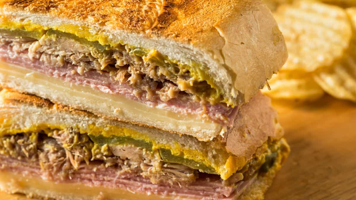 A close-up of a cuban sandwich with layers of meat, cheese, and pickles, accompanied by potato chips on the side.