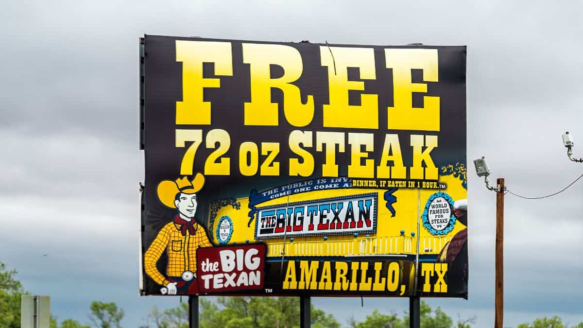 Billboard advertising a free 72 oz steak challenge at the big texan in amarillo, texas, featuring retro comic-style graphics under a cloudy sky.