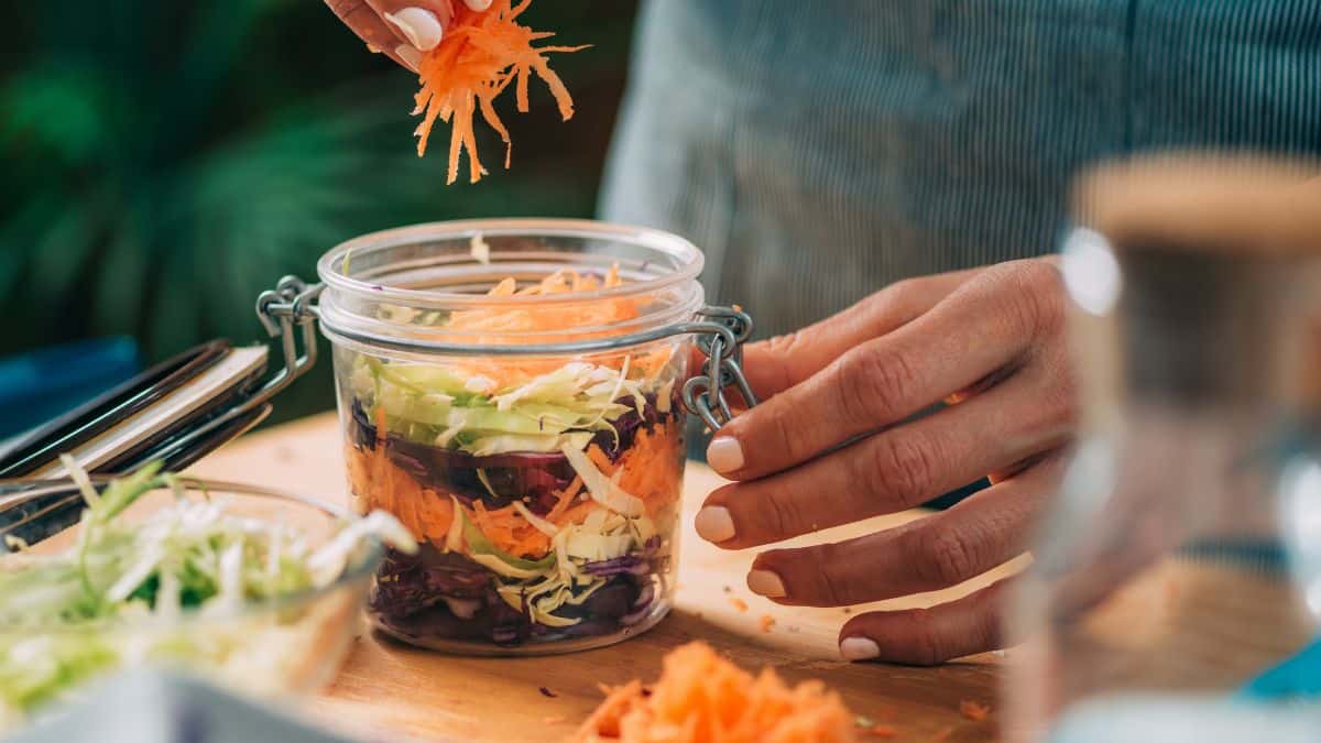 A person preparing a layered salad by adding shredded carrots to a mason jar.