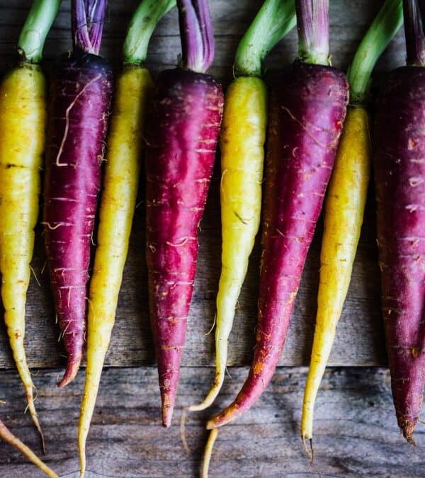 Colorful carrots in yellow, red, and purple, with green tops, aligned on a rustic wooden surface.