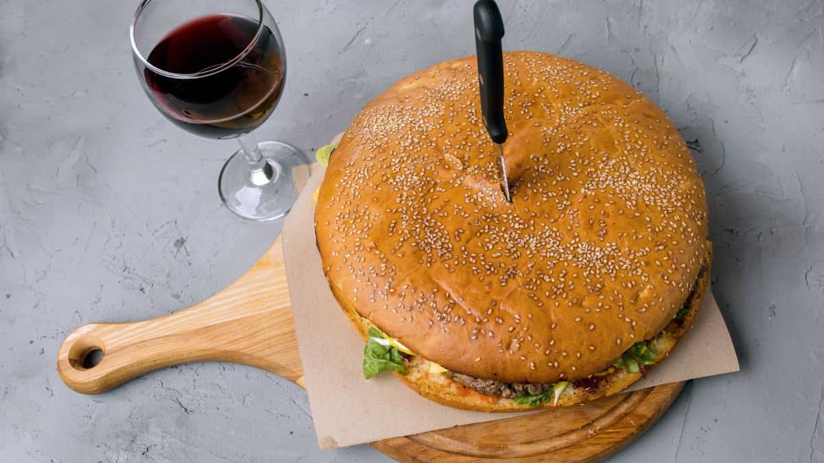 A large hamburger with a knife in the center on a wooden board, accompanied by a glass of red wine on a gray surface.