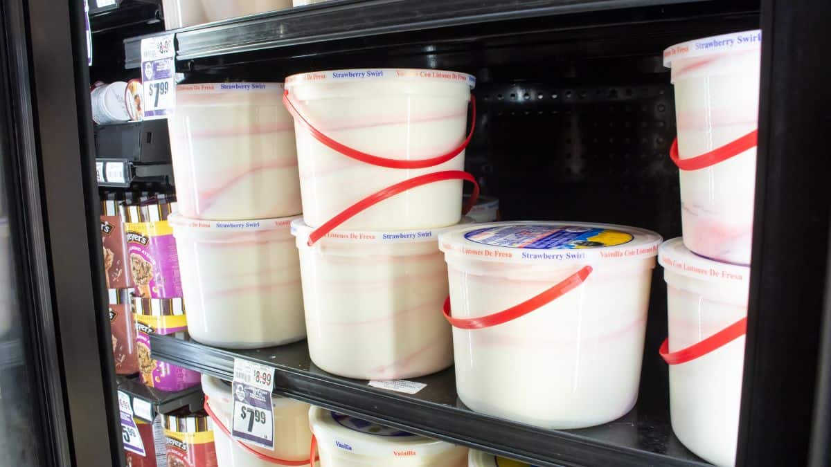 Open freezer displaying various large tubs of strawberry swirl ice cream secured with red rubber bands, alongside other frozen desserts.