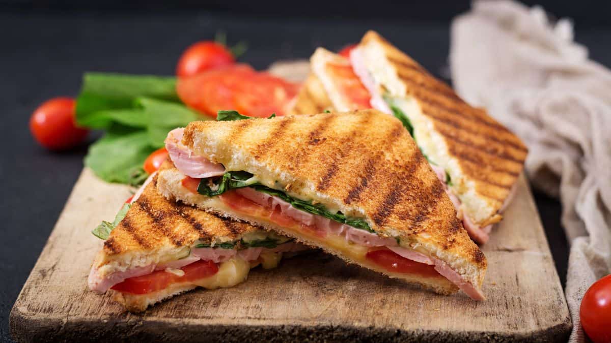 Grilled sandwich with ham, cheese, tomato, and lettuce on a wooden cutting board.