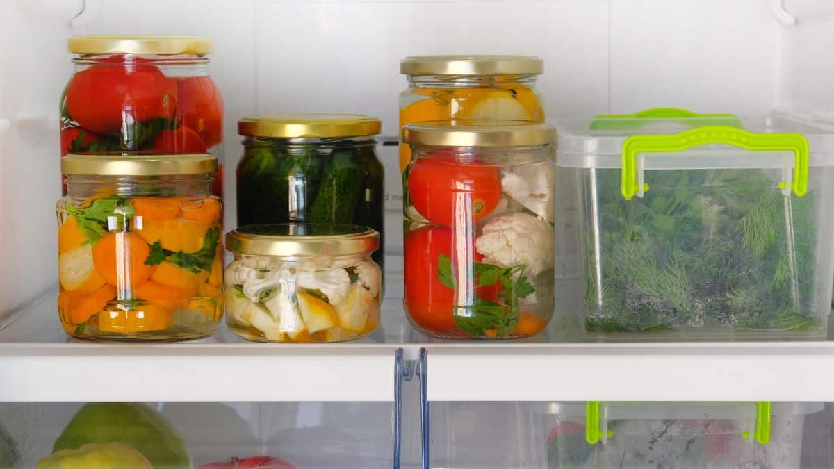Assorted pickled vegetables in jars on a refrigerator shelf.